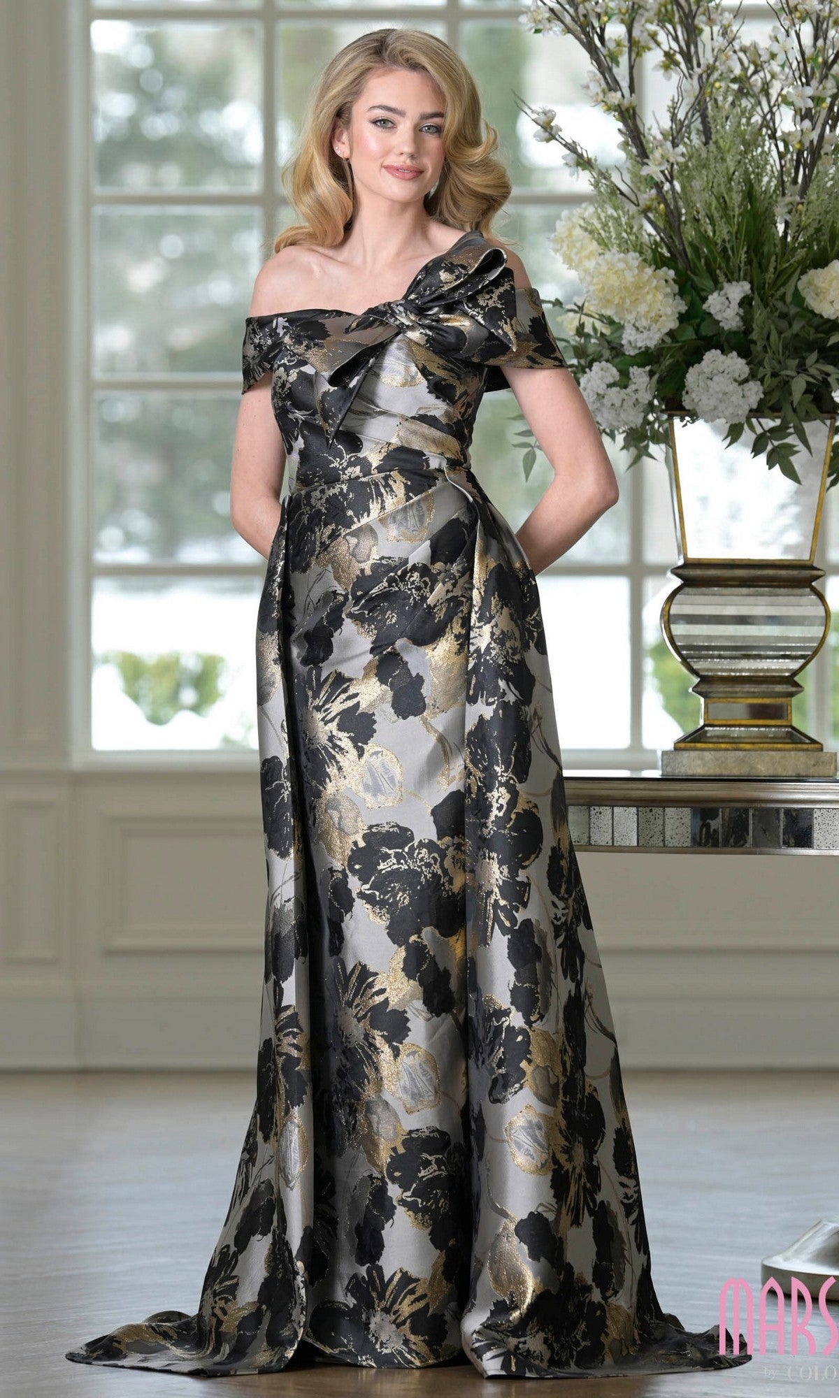 A woman stands indoors near a large floral arrangement, wearing the Marsoni MV1396 Long Formal Dress in Black Multi—an off-the-shoulder, floor-length jacquard gown with black, gold, and silver floral accents.